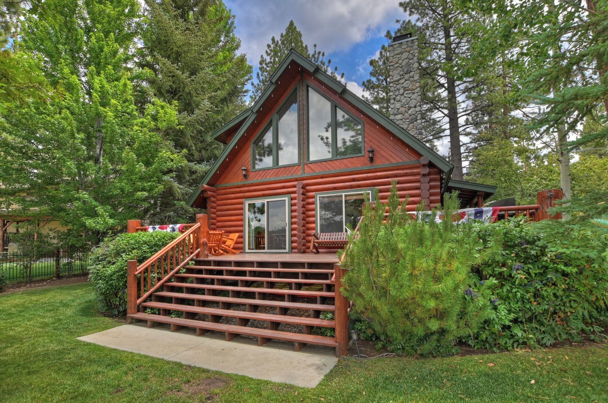 A log cabin with a wooden staircase leading to a deck, surrounded by lush greenery.