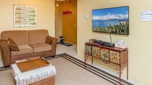 Ein Wohnzimmer mit braunem Sofa, einem Couchtisch, einem Fernseher auf einem Holzständer, einem Bilderrahmen mit einem Seeblick und einem Schild mit der Aufschrift 'LAKE RULES'.