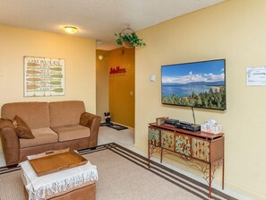 Uma sala de estar com um sofá marrom, uma mesa de centro, uma TV sobre um suporte de madeira, um quadro com uma paisagem de lago e uma placa com a inscrição 'REGRAS DO LAGO'.