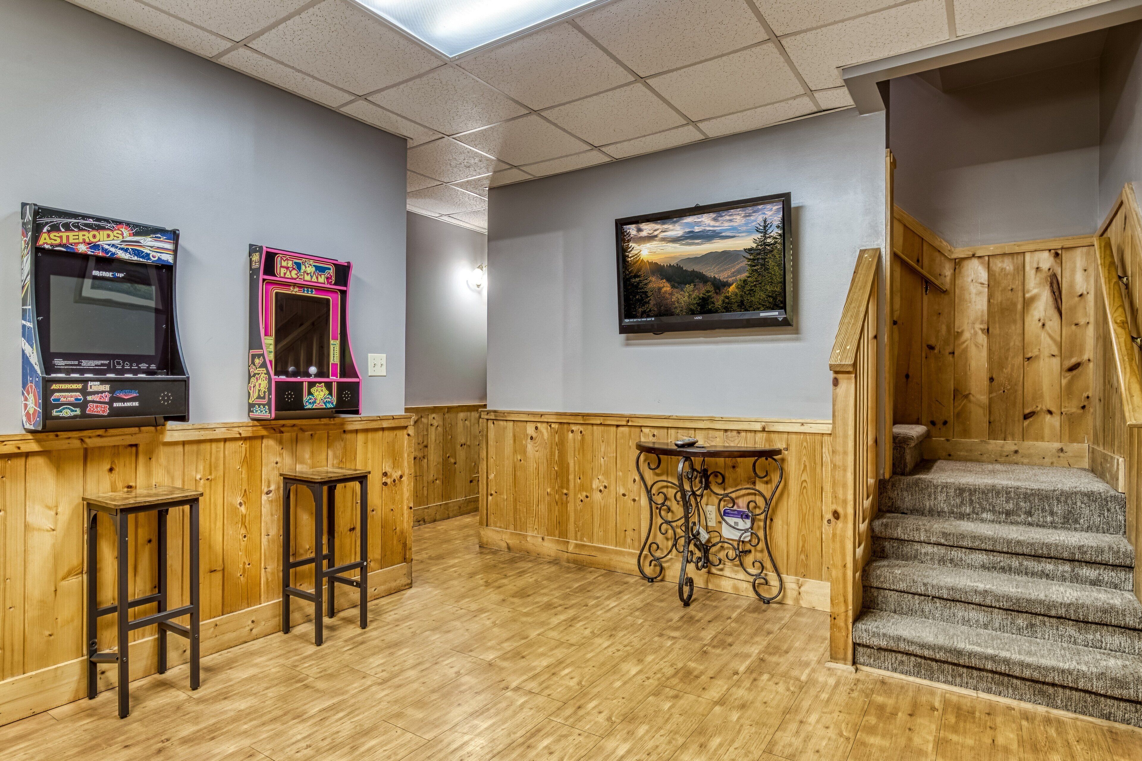 A bar area with wooden paneling, two arcade games, a small table, and a staircase.