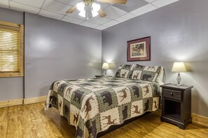 Un dormitorio con piso de madera, una cama con colcha, dos lámparas de noche, una mesita de noche, un ventilador de techo y un cuadro en la pared.