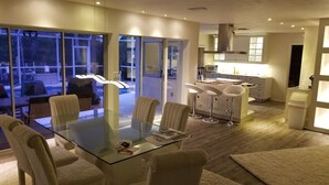 A modern dining area with a glass table and white chairs, a kitchen with white cabinets, and a view through large windows.