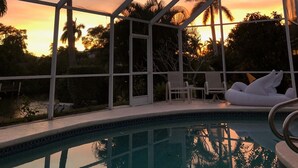 A poolside area with a glass enclosure, lounge chairs, and a view of palm trees and sunset.