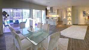 A spacious living area with a glass-top dining table, white sofas, and a kitchen in the background.