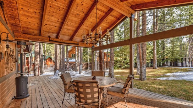 A wooden deck with a round table and chairs, surrounded by tall trees. - Running Bear by Destination Big Bear : Walk to Snow Summit! Pool Table!Great Backyard w/ Hot Tub! Master Suite! (Big Bear Lake)