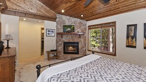 A bedroom with a stone fireplace, a bed with a patterned bedspread, a ceiling fan, and a window with a view of trees. - Running Bear by Destination Big Bear : Walk to Snow Summit! Pool Table!Great Backyard w/ Hot Tub! Master Suite! (Big Bear Lake)