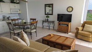 A living room with a sofa, a coffee table, a TV, a dining table with chairs, a kitchen area with a microwave, and a clock on the wall.