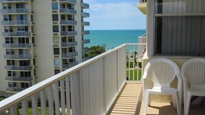 A balcony with white railings, two white plastic chairs, and a view of the ocean and adjacent high-rise buildings.