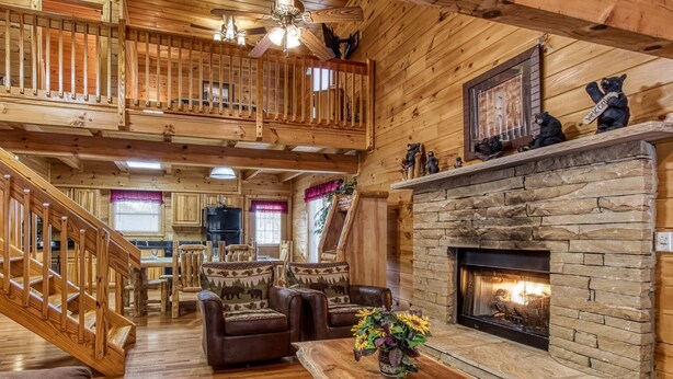 A wooden cabin interior with a stone fireplace, wooden staircase, and a dining area. - Point Of View - Spacious 2 Bedroom With a Loft Just off Glades Road (Gatlinburg)