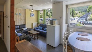 Een gezellige zithoek met een bank, een salontafel en een koelkast. Er is een raam met uitzicht op geparkeerde auto's en een bergachtig landschap.