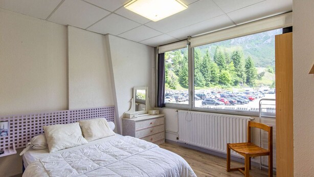 Een slaapkamer met een bed, een houten stoel, een nachtkastje, een spiegel en een raam met uitzicht op een parkeerplaats en bomen.