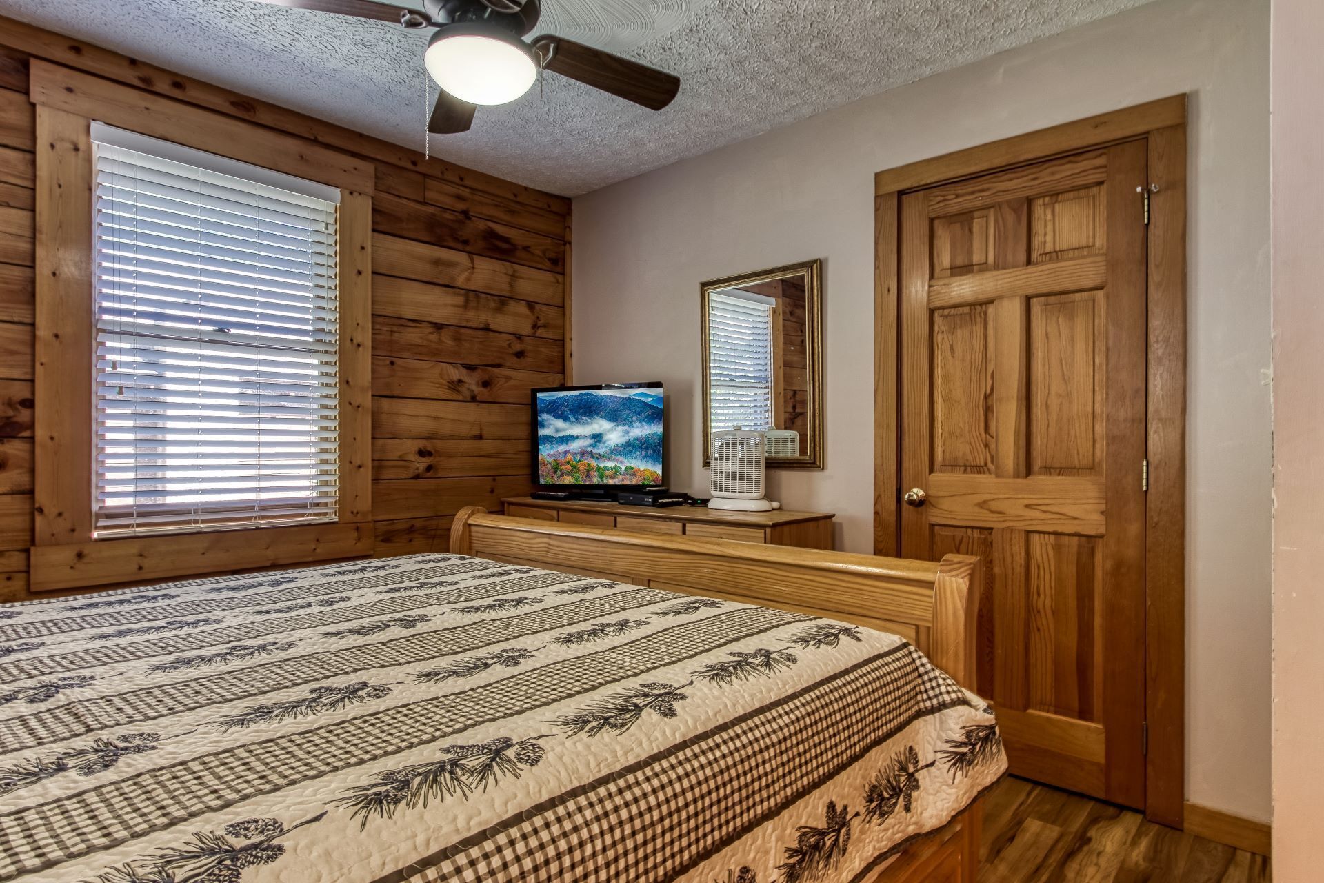 A bedroom with a wooden ceiling, a ceiling fan, a bed with a patterned bedspread, a television on a wooden dresser, and a door with a mirror.