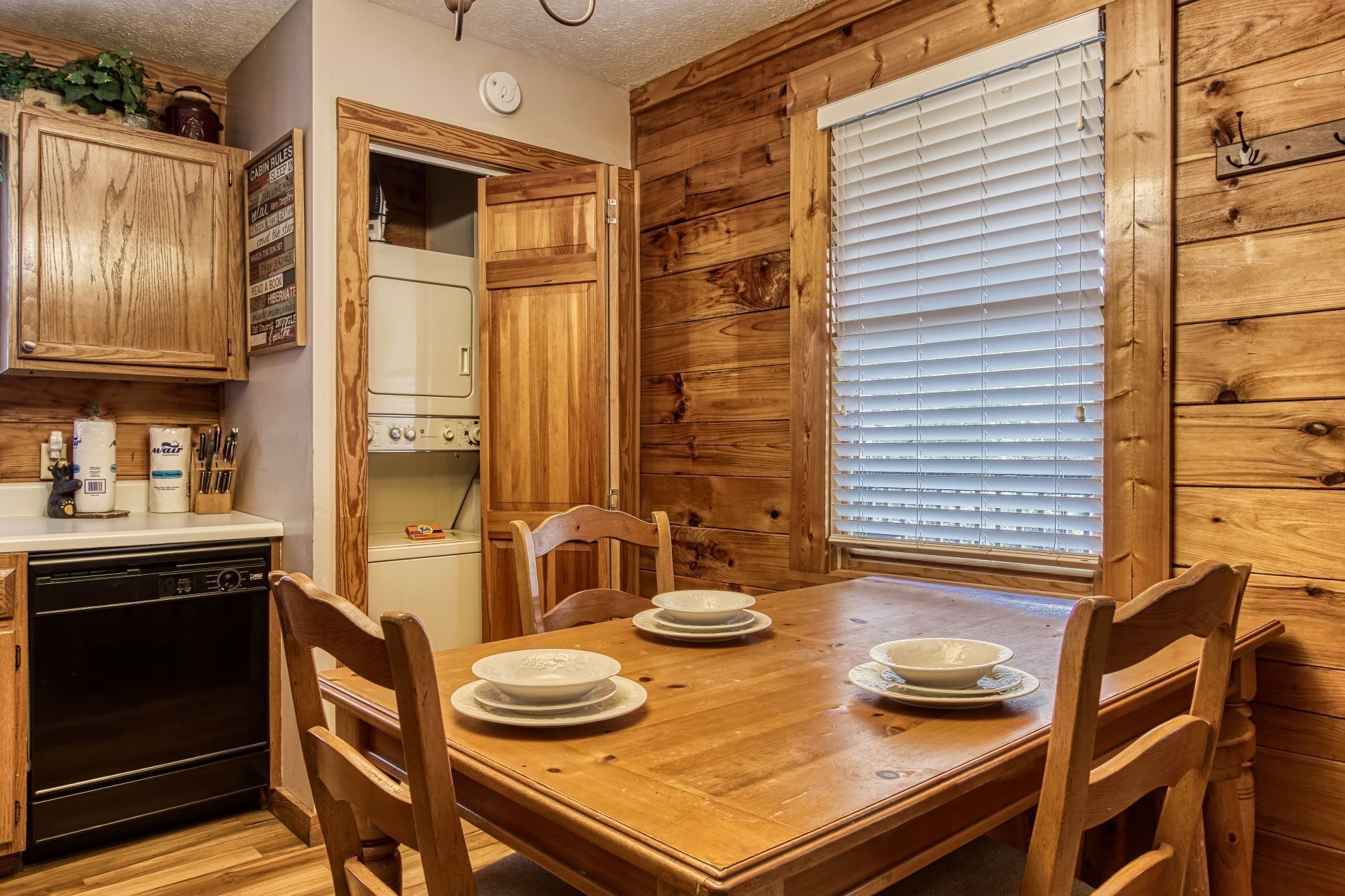 A wooden cabin interior with a dining table set for four, a washing machine, and a kitchen area with wooden cabinets.