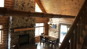 A log cabin interior with a stone fireplace, wooden beams, and a dining area. - The Driftless Escape! Modern, large cabin/57 private acres in WI drift less area (Richland Center)