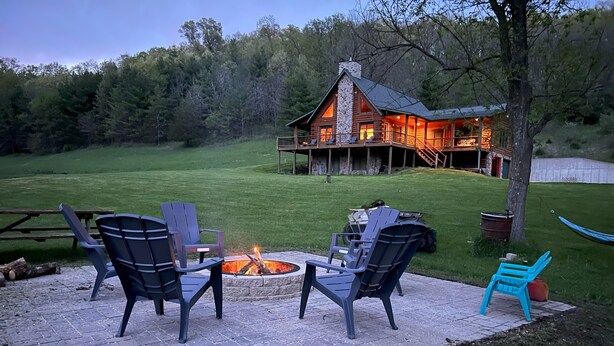 A cozy outdoor seating area with a fire pit, surrounded by stone tiles, and a wooden deck leading to a rustic cabin with a stone chimney. - The Driftless Escape! Modern, large cabin/57 private acres in WI drift less area (Richland Center)