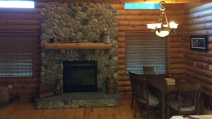 A log cabin interior with a stone fireplace, wooden ceiling beams, and a dining area with a table and chairs. - The Driftless Escape! Modern, large cabin/57 private acres in WI drift less area (Richland Center)