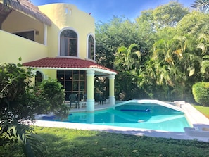 Una piscina con terraza blanca y zona cubierta con sillas.