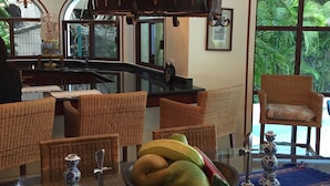 Een eethoek met rieten stoelen, een glazen tafel met een schaal fruit, en een kroonluchter met meerdere lampen.