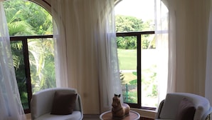 Een kamer met twee rieten stoelen, een klein rond tafeltje met een decoratief paardbeeld, en grote ramen met lichte gordijnen.