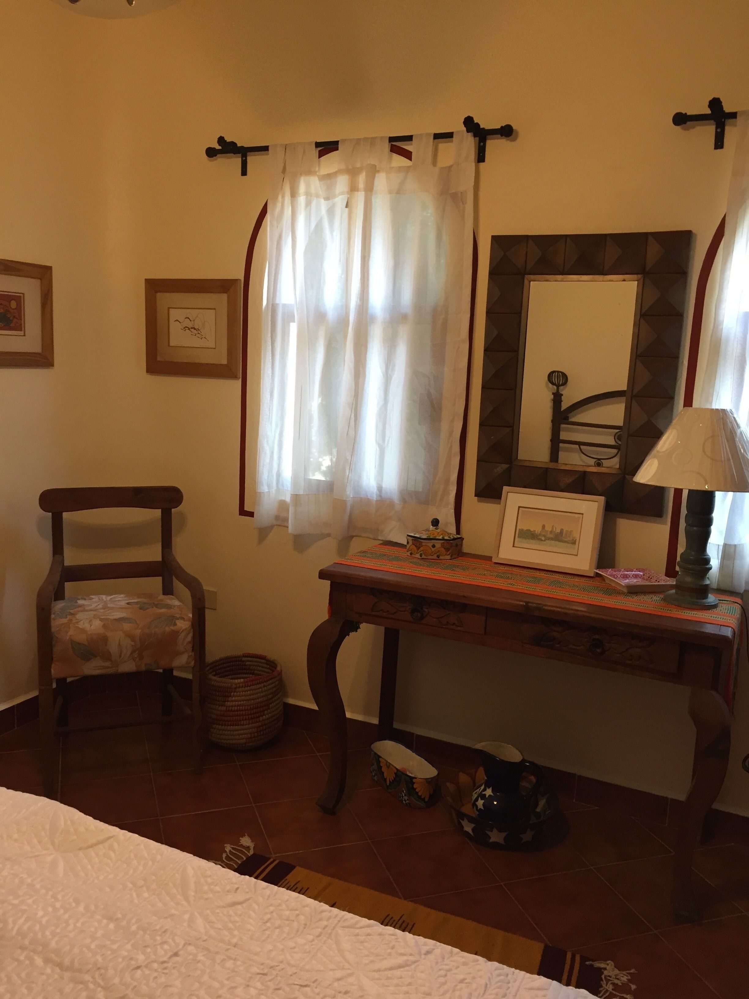 Ein Zimmer mit einem Bett, einem Holzstuhl, einem kleinen Tisch mit Lampe, einem Spiegel und einem Fenster mit Vorhängen.