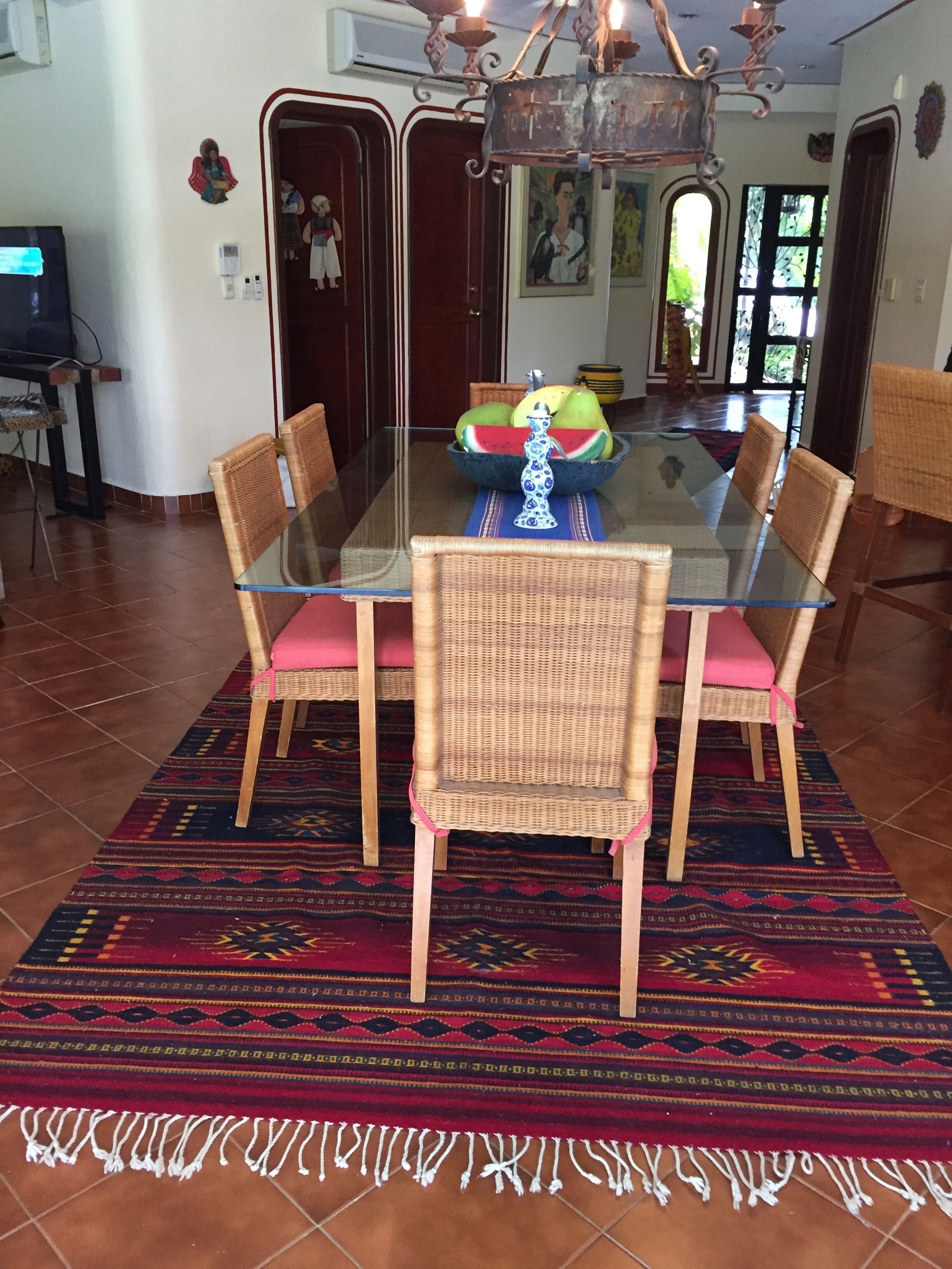 Un comedor con mesa de vidrio, sillas de mimbre con cojines rosa y un tapete tejido colorido.