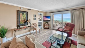 A spacious living room with a large window offering a view of the ocean, a comfortable seating area with a red armchair, and a flat-screen TV mounted on the wall. - 🌊 Islander #508 – Beachfront Bliss with Amazing Views! 🏖️✨ (Destin)