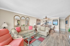 A spacious living room with a glass coffee table, two sofas, a dining area with a chandelier, and a large mirror on the wall.