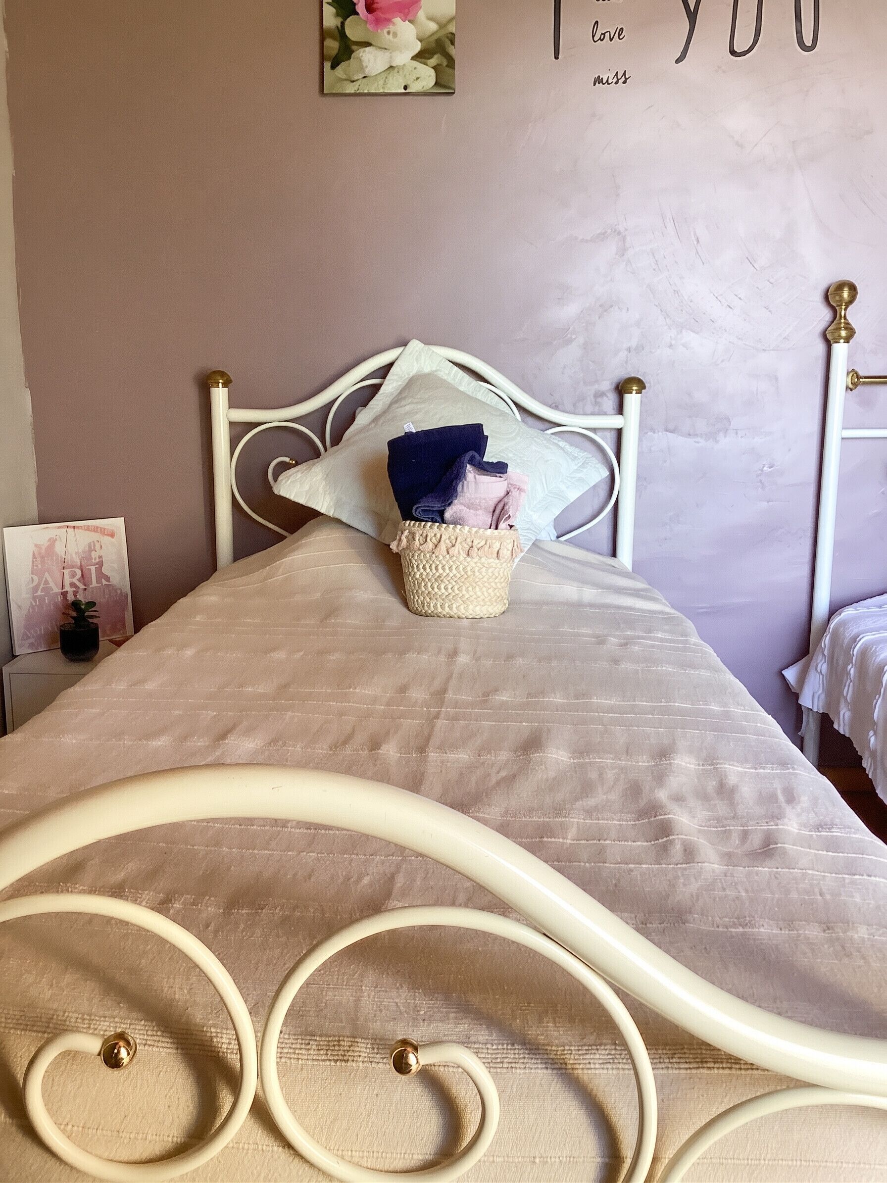 Een bed met een beige sprei, een geweven mand met opgevouwen kleding, en een muur met een 'love you miss' bord.'