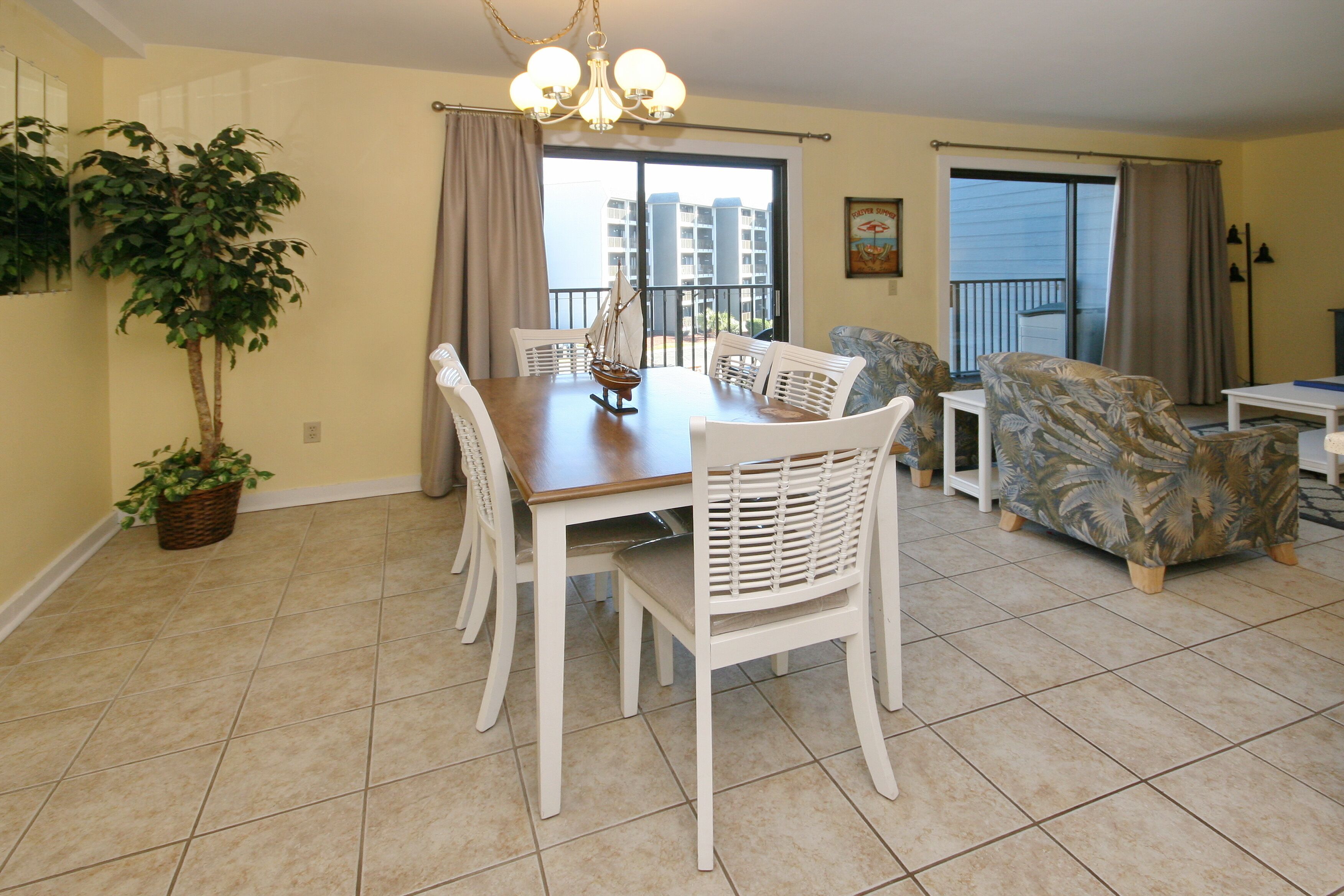 Un coin repas avec une table en bois et des chaises blanches, une plante en pot et une vue sur un balcon doté d’une balustrade.