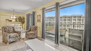 Un salon spacieux avec deux fauteuils à motifs, une table basse blanche et un coin repas avec une table et des chaises blanches. Une grande fenêtre offre une vue sur un immeuble et sur du mobilier de balcon.