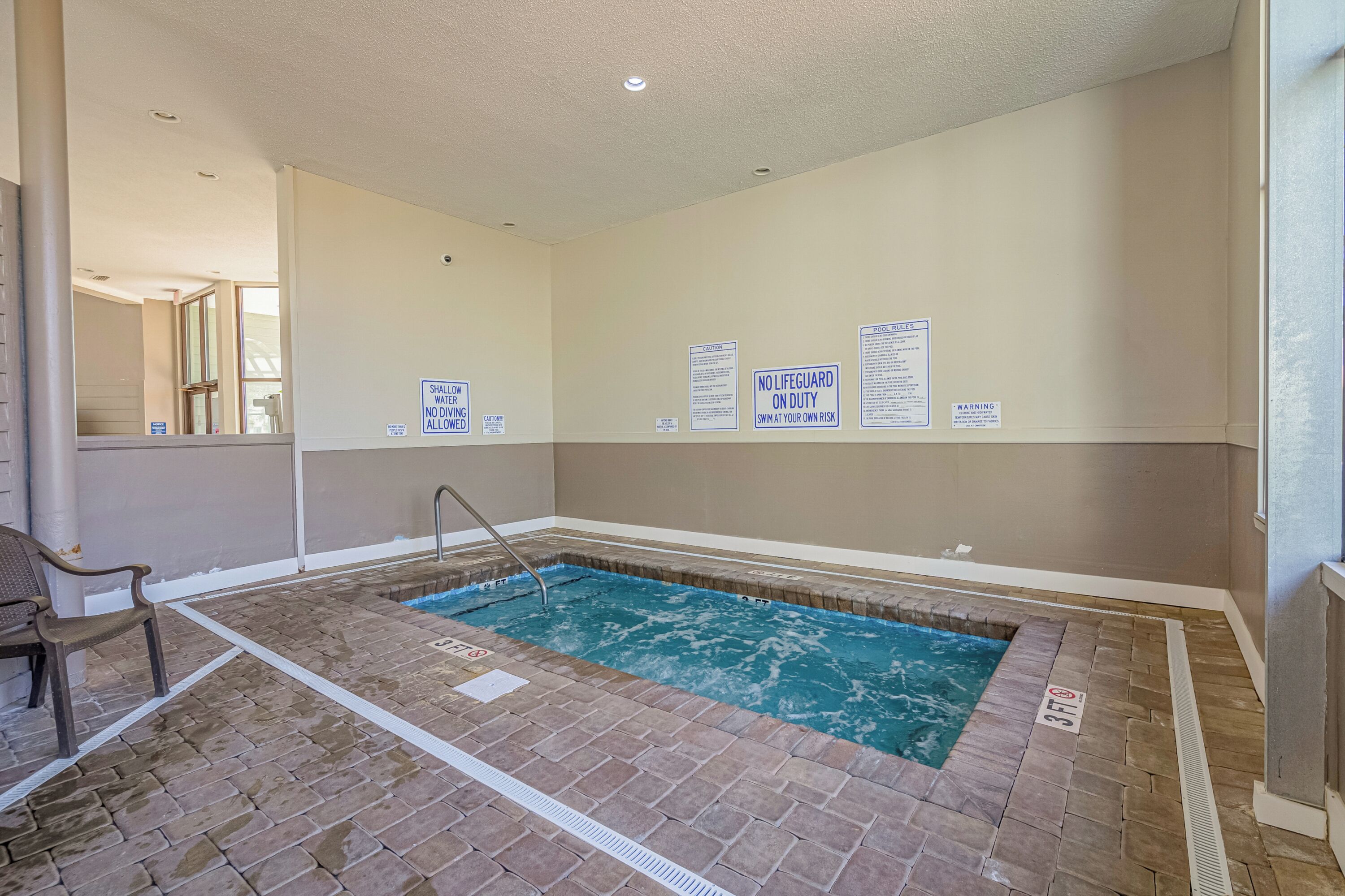 An indoor swimming pool with safety signs, a brick floor, and a chair.