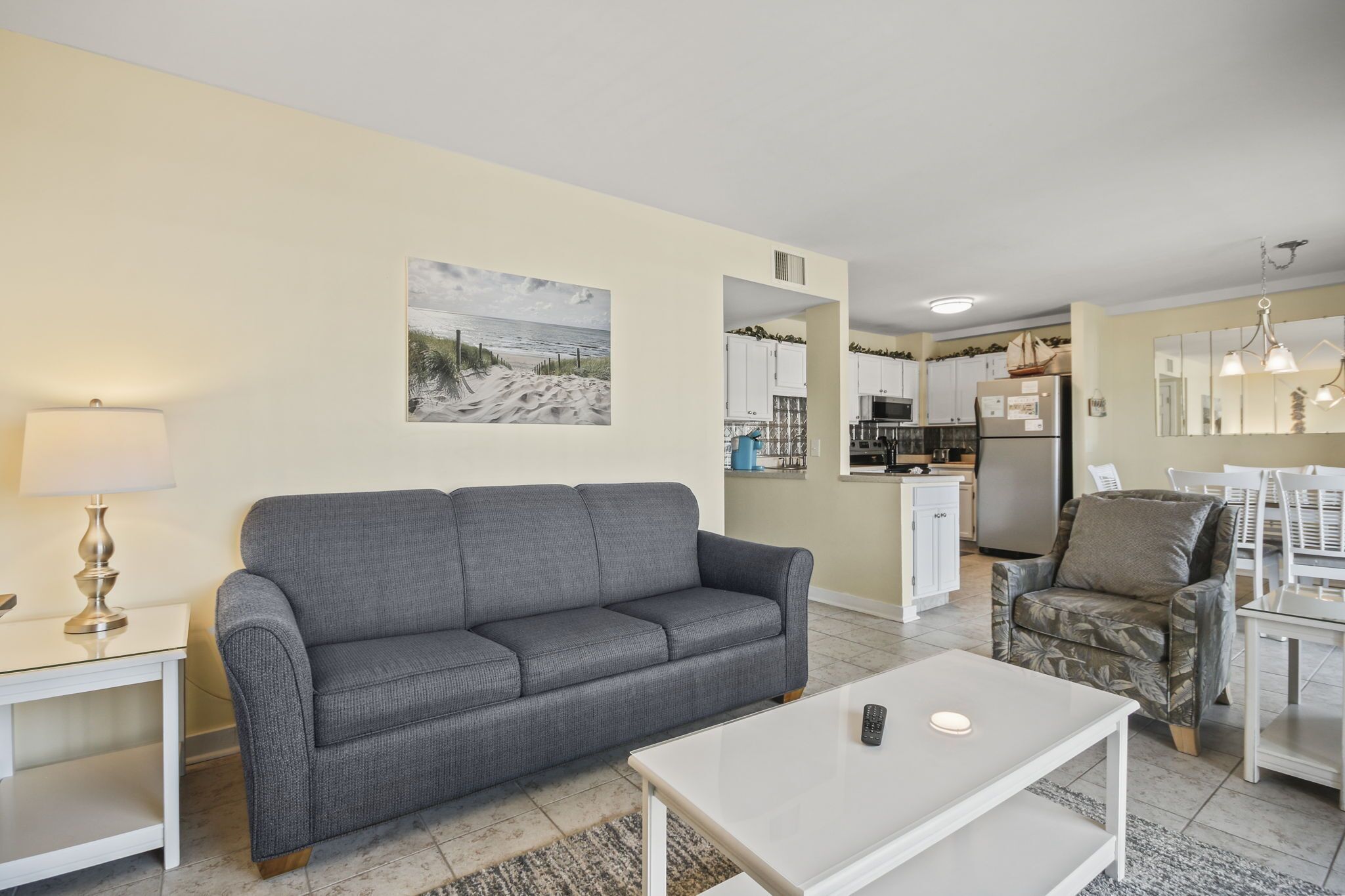 A living room with a grey sofa, a white coffee table, and a lamp. There is a kitchen area with white cabinets and a refrigerator.
