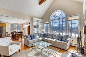 A spacious living room with a high ceiling, a large window offering a mountain view, a dining area with wooden furniture, and a kitchen with a stainless steel refrigerator.