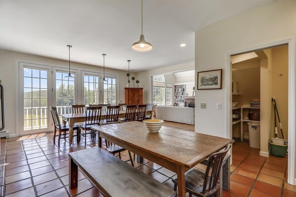A spacious dining area with a large wooden table and chairs, a kitchen in the background, and a view of the outdoors through large windows.