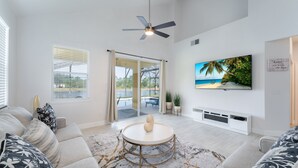 A modern living room with a ceiling fan, a large TV on the wall, a round table with a decorative object, and a view of the outdoors through a sliding glass door. - Lake View! Amazing Sunsets w/West Facing Pool (Clermont)
