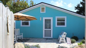 Une petite maison bleue avec une porte blanche, deux fenêtres et une véranda avec une chaise et un parasol.