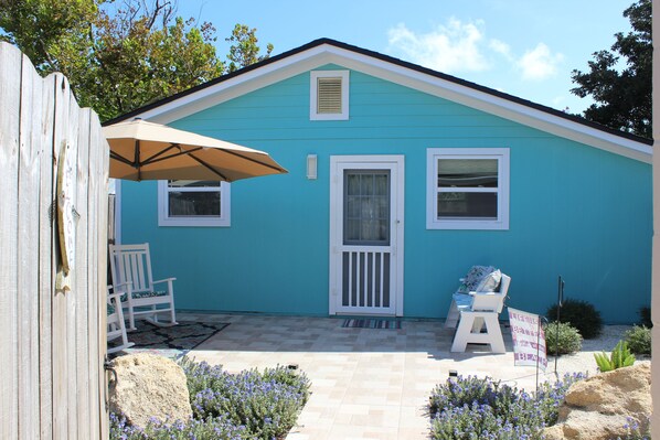 A small blue house with a white door, two windows, and a porch with a chair and an umbrella.