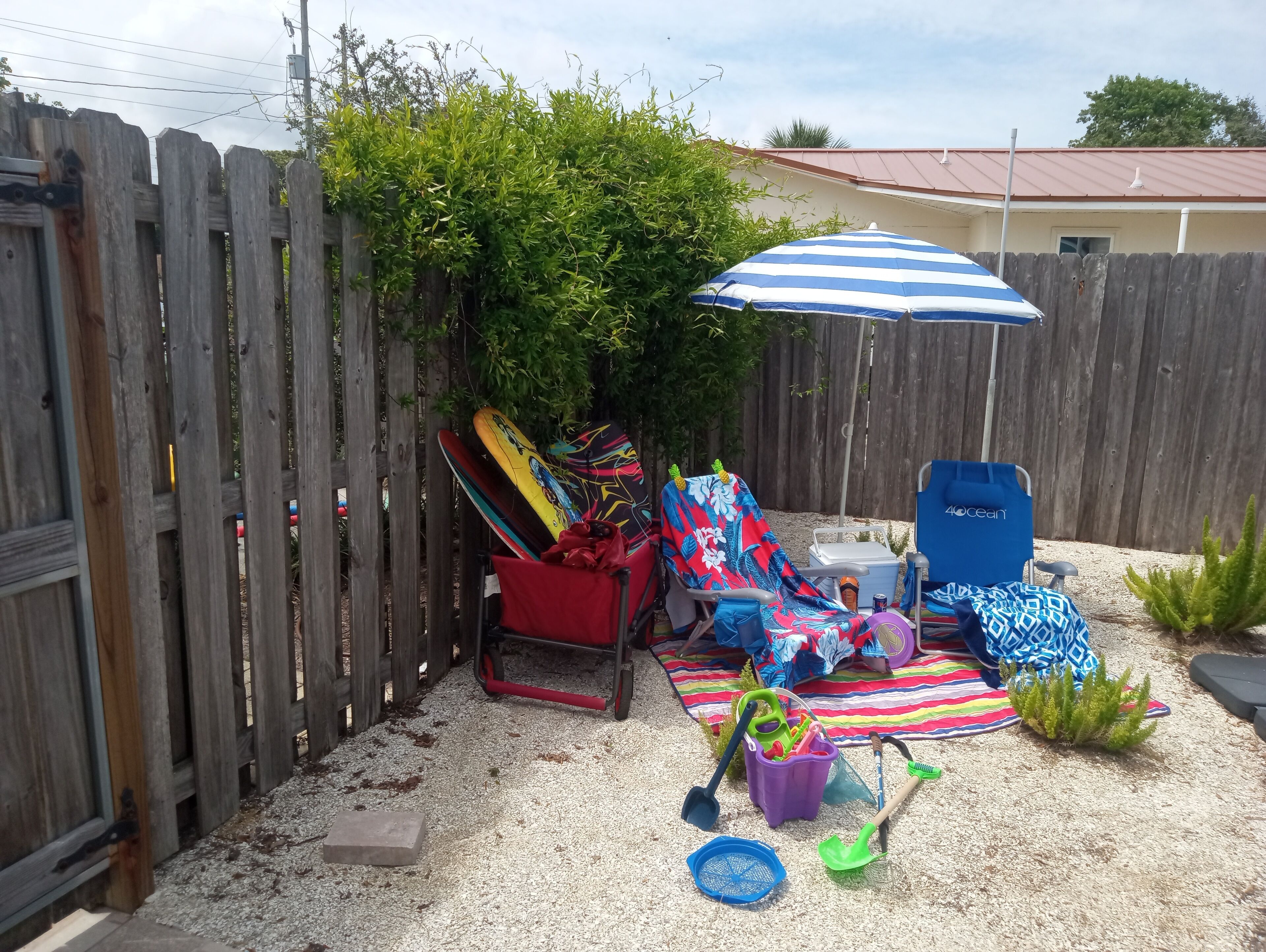 A backyard with a wooden fence, a covered seating area with a striped umbrella, and various beach toys.