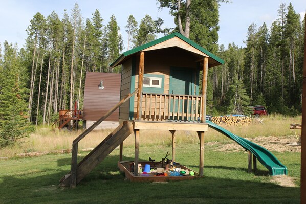Une cabane en bois avec un toboggan, entourée d’une pelouse et d’arbres.