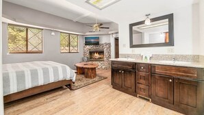 Un dormitorio con una cama, chimenea de piedra, una mesa redonda de madera y un gran ventanal con persianas.
