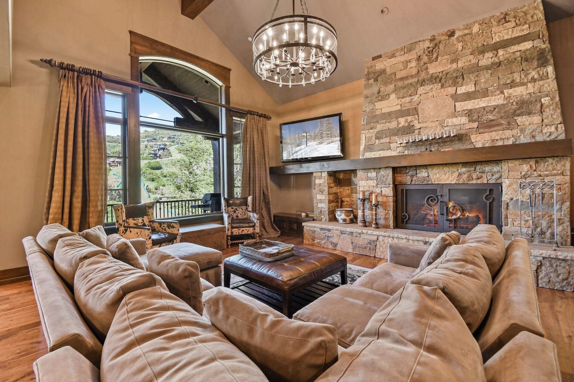 A living room with a stone fireplace, leather sofas, a chandelier, and a large window with curtains.