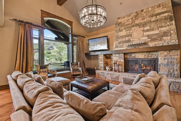 A living room with a stone fireplace, leather sofas, a chandelier, and a large window with curtains.