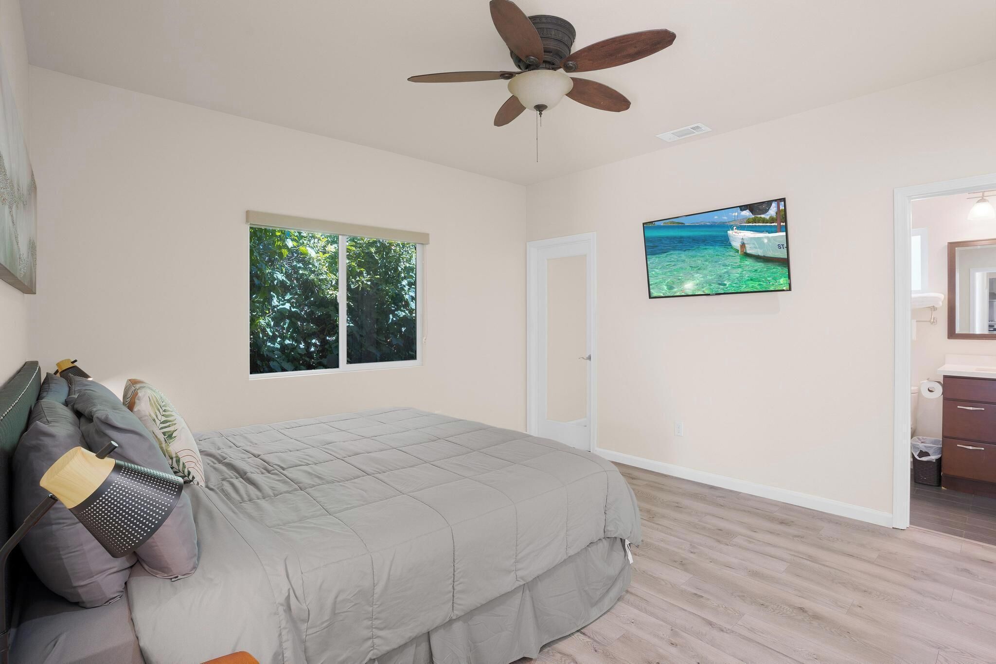 A bedroom with a bed, a ceiling fan, a window with a view of trees, a door, and a TV on the wall.