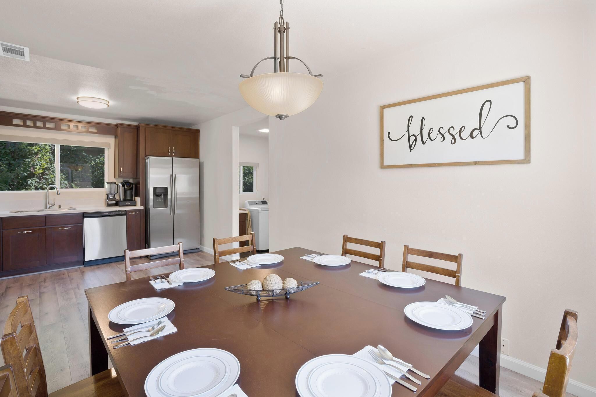 A dining area with a wooden table set for four, surrounded by chairs, with a kitchen in the background.