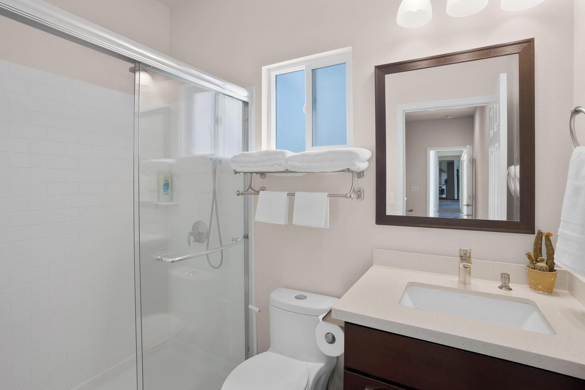 Un baño moderno con cabina de ducha de vidrio, inodoro blanco, lavamanos con espejo y ventana con vistas al exterior.