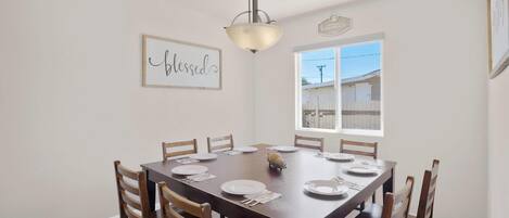 Comedor con una mesa de madera dispuesta para seis comensales, una araña y una ventana que ofrece vistas a una casa.