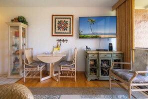 A room with a round table, two chairs, a cabinet with glass doors, a framed floral artwork, a wall-mounted TV displaying a beach scene, and a woven rug.