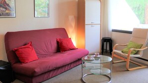 A red sofa with two red cushions, a glass coffee table with a cup and saucer, a white chair with a green cushion, and a white cabinet with two drawers. - Charming Studio in Vars, 30m from Slopes, Sleeps 4 (Vars)