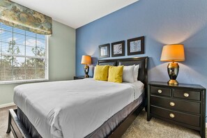 Une chambre à coucher avec un lit, deux tables de chevet avec des lampes, une fenêtre avec des stores et trois tableaux encadrés accrochés au mur.