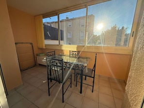 A small dining area with a glass table and chairs, a window with a view of buildings, and a closed shutter door.